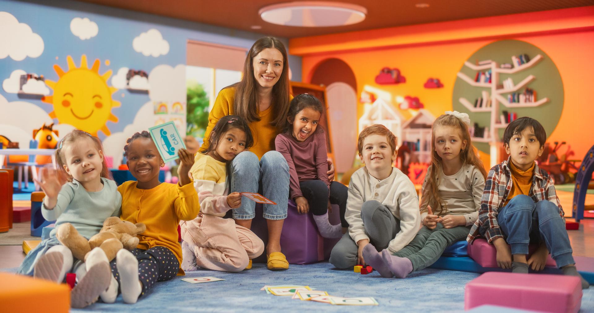 Portrait of Happy Diverse Children Posing Together with a Female Preschool Teacher in a Modern Kindergarten. Educator and Beautiful Small Kids Looking at Camera, Smiling, Waving Hands