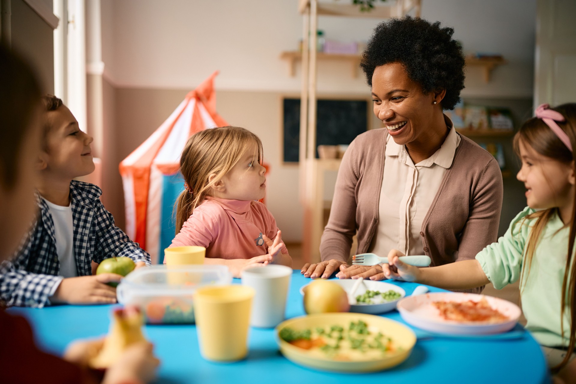 Happy black teacher talking to group of kids during lunch time at preschool.